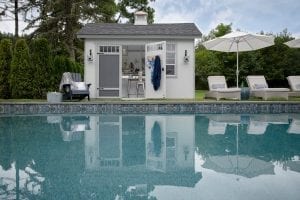 Pool shed with one door open next to in-ground pool and chairs.