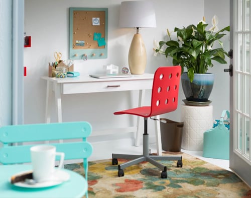 Home office She Shed: Desk and lamp, red swivel chair, colorful rug, and cork board.